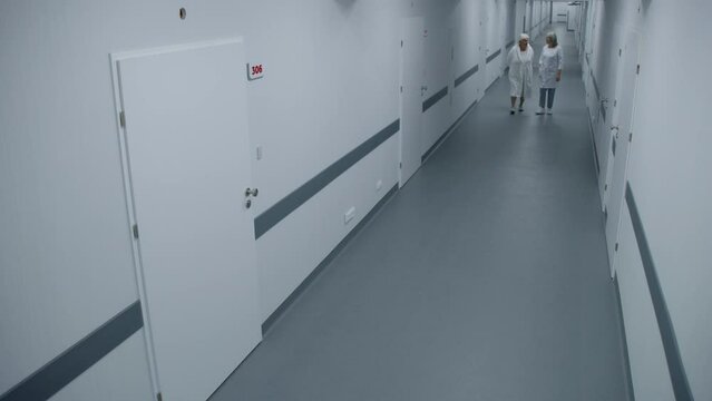 Bright clinic corridor: Female doctor and elderly woman walk down the hallway to hospital ward, talk about treatment. Medical staff work in modern medical facility. Security camera view. High angle.