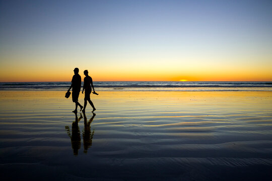 Young couple in love, unidentifiable, silhoutted by the setting sun, walking at the edge of the sea at Cable Beach, Broome, Western Australia. 