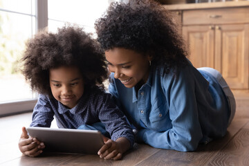 Close up caring African American mother with little daughter using tablet, looking at screen, relaxing lying on wooden floor at home together, smiling mom with 7s girl child spending leisure time