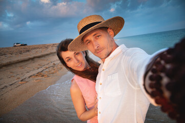 Close-up of smiling happy young couple, two family man and woman hugging each other, taking first person selfie at sunrise or sunset on sea beach on summer day