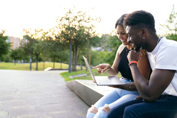 A charming couple of different ethnicities, urban chic, smiles, and laptop, captivated by the sunset's glow in the city park