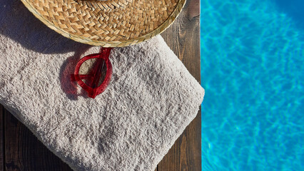 16:9 still life relaxation concept on vacation. Summer accessories next to a pool of blue water. Towel, sunglasses and straw hat.