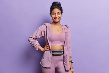 Attractive Iranian sporty woman with dark hair keeps hand on waist stands satisfied dressed in sportswear smiles gladfully isolated over purple background leads healthy lifestyle. Sport is life - 648807525