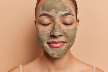 Headshot of young Latin woman applies facial clay mask has eyes closed undergoes beauty procedures for refreshing skin stands bare shouldered isolated over brown studio background. Cropped image