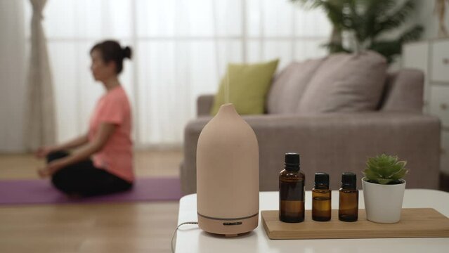 closeup of steam coming out from aromatherapy vaporizer electronic device and essential oil bottles with asian female adult sitting in meditative pose on background
