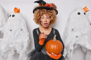 Indoor photo of young surprised African american female in costume of witch holding pumpkin...