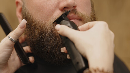A barber cuts a bearded man's hair with a clipper in a barbershop. Men's hairstyle and haircut in...