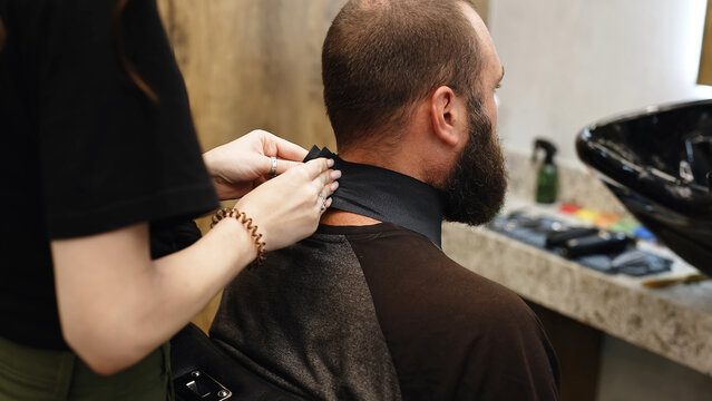 The photo shows a young stylish female hairdresser putting a cape on a client. Fashionable hairdresser at work in a retro barbershop. Preparing for a haircut and grooming for a stylish man.