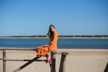 Pretty young blonde woman in orange dress sitting on the wooden railing of the beach promenade. In...