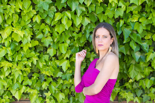 Pretty young blonde woman in purple dress posing for photos against background of a wall of ivy. Woman makes different expressions, serious, happy.