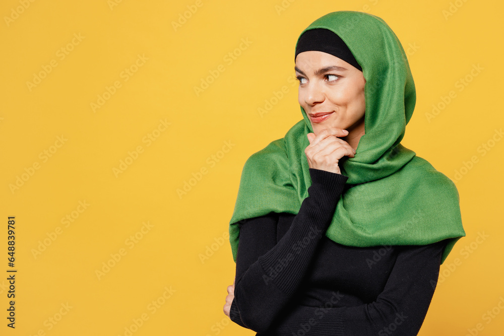 Wall mural Young happy arabian asian muslim woman wear green hijab abaya black clothes put hand prop up on chin look aside isolated on plain yellow background. People uae middle eastern islam religious concept.