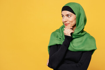 Young happy arabian asian muslim woman wear green hijab abaya black clothes put hand prop up on chin look aside isolated on plain yellow background. People uae middle eastern islam religious concept.