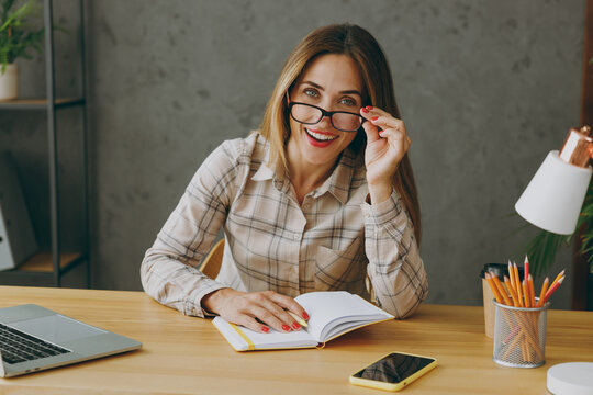 Young Successful Employee Business Woman Wear Shirt Casual Clothes Touch Glasses Sit Work At Office Desk With Pc Laptop, Look Camera Write Down In Notebook, Take Notes. Achievement Career Job Concept.