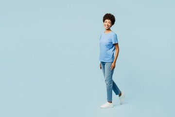 Full body side profile view young fun woman of African American ethnicity wear t-shirt casual clothes walking going look camera isolated on plain pastel light blue cyan background. Lifestyle concept.