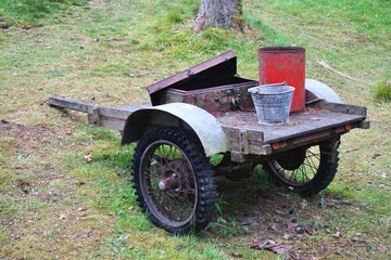 Vintage homebuilt trailer with spoked knobby tread wheels hauling a galvanized bucket, square metal box with open lid and tall red painted tin container