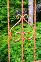 Vintage rusted iron fence with a single heart-shaped brass padlock hanging from an ornamental scroll with blurred bushes in the background