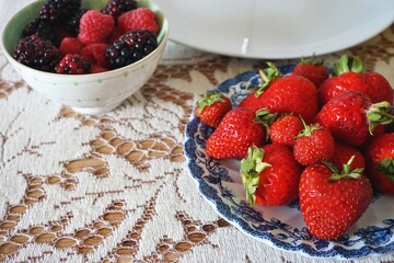 Fresh ripe berries on ceramic plate and bowl on top of white lace tablecloth with copy space at one corner