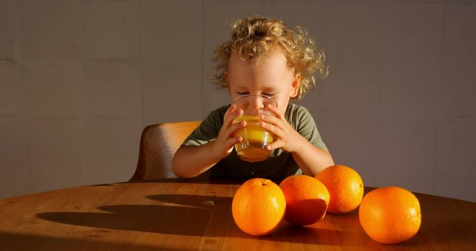 Young child takes glass of orange juice and makes very small sip, squinting slightly, apparently because juice is sweet and sour. Then he places glass on table beside big, bright oranges.