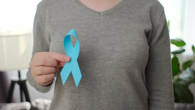 Symbol for support men. Close-up of female hand demonstrating small blue ribbon standing in living room. Selective focus. Healthcare, International men, Father and World cancer day concept