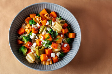 Autumn pasta salad with roasted pumpkin, broccoli, feta cheese on dark wooden background, vivid colors fall season salad, autumn food concept, top view