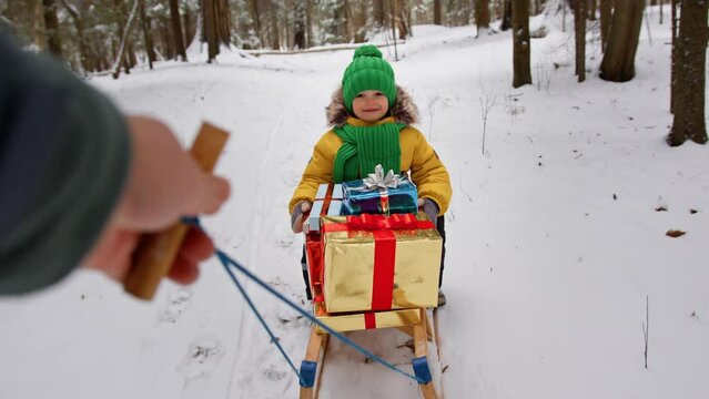 Preschooler in bright winter jacket holding bunch of New Year presents on sleigh