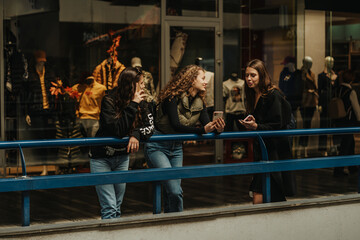 Three girls leaning on a blue fence. Curly haired girl showing something on the phone to her friend while their long brunette friend is having a phone call and looking at them