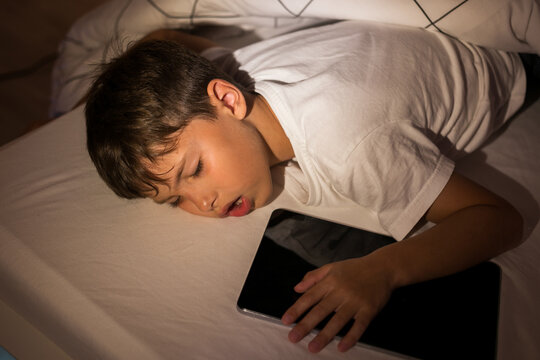 Little school-age boy fell asleep on the bed playing games on the tablet, hugging the gadget, sleeping. Child sleeps in his bed in the children's room. At night or in the evening he fell asleep.