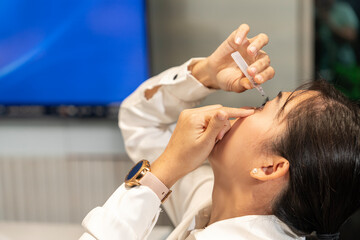 Obraz na płótnie Canvas Young female Asian employee fingers her eyes and applies artificial tears or eye drops to the eyeballs to reduce eye strain while sitting at a desk in the company.