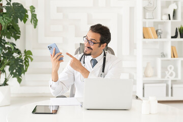 Close up portrait of smiling male doctor sitting at the desktop and talking to phone in the office...