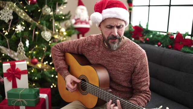 Young bald man singing song playing classical guitar sitting by christmas tree at home