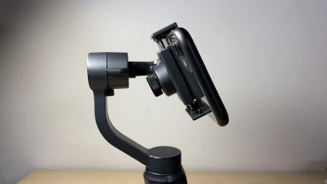 Black automatic gimbal stabilizer holding black phone on table against white wall indoor. Technology and electronic gadget concept 