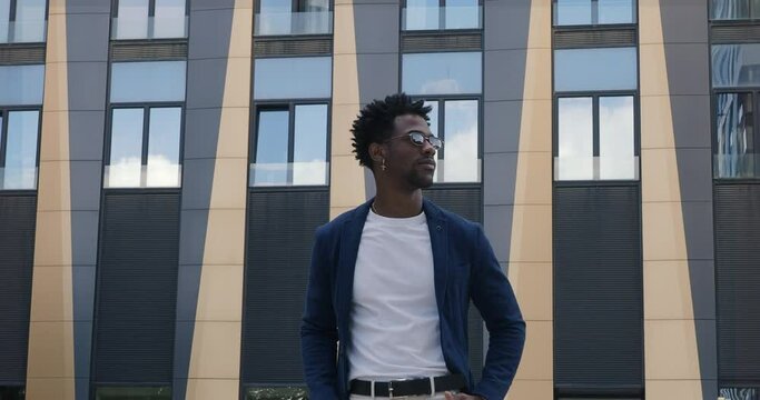 Handsome african American guy in stylish clothes stands in city next to modern building, looking to side. Portrait stylish black man