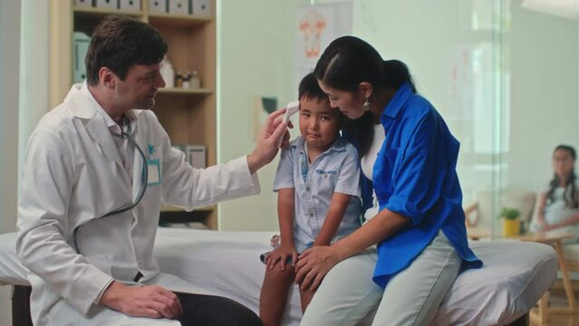 Medium long shot of male general practitioner measuring body temperature of little boy with infrared thermometer during annual check-up in clinic