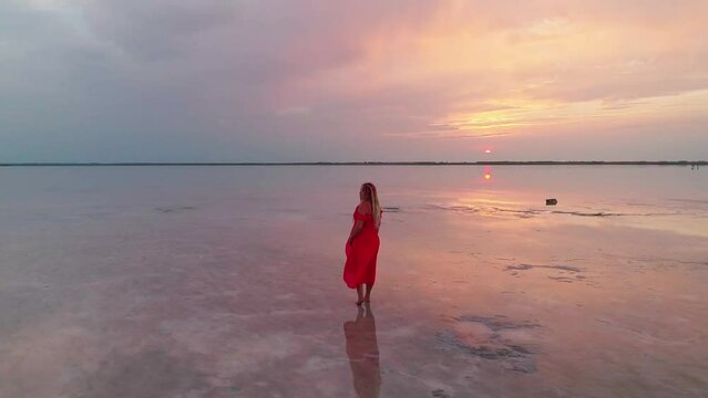 Aerial of a young woman in red dress standing in the water of a unique pink salt lake. Sunset at lake Bursol with beautiful reflections on calm water surface. Stunning scenery
