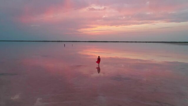 Aerial of a young woman in red dress walking in the water of a unique pink salt lake. Sunset at lake Bursol with beautiful reflections on calm water surface. Stunning scenery