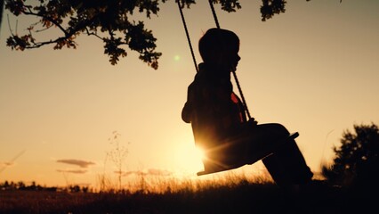 Child plays on wooden swing, dreams of fly. Happy little boy swings on swing in park under tree at...