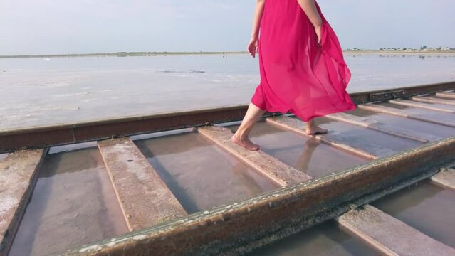 Young woman in red dress walking on a railway in the water of a unique pink salt lake Bursol. Unique scenery