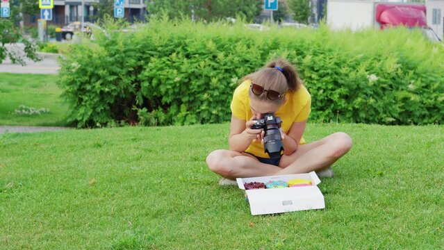 Female food photographer sits on grass, takes pictures of donuts inside paper packaging. Close up. Girl shooting doughnuts with professional camera in box on lawn for her blog about nutrition, diets