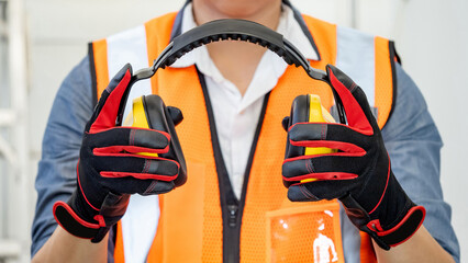 Male worker with reflective orange vest and protective hand gloves pulling yellow safety ear muffs...