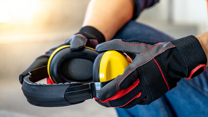 Male worker hand with black and red protective gloves holding yellow safety ear muffs or ear protectors preparing to wear on his head. Equipment for high noise reduction - obrazy, fototapety, plakaty