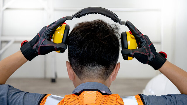Back view of male worker with reflective orange vest and protective hand gloves pulling yellow safety ear muffs or ear protectors when wearing on his head. Equipment for high noise reduction