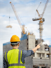 Asian male construction worker or site engineer man with green reflective vest and yellow safety helmet pointing at unfinished building structure and tower crane