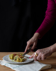 A lady whose hands can only be seen delicately cuts a stuffed cabbage roulade. A white stone ring on her finger and purple nail varnish on her nails. Dark mood, black background and copy space.