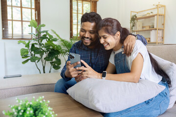 happy indian couple relax sitting on sofa at home holding phone video calling distance with family