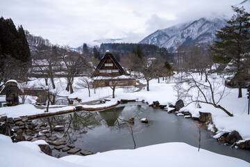 Fototapeta na wymiar In the tranquil setting of Shirakawa village, the reflection of a snow-thatched roof cottage is beautifully mirrored in a small pond.