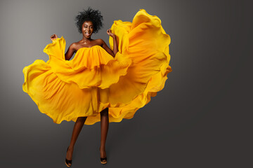 Happy smiling Woman in Orange Dress flowing on Wind over Gray. Fashion African Model with Afro Curly Hair in yellow Chiffon Gown. Cheerful Girl Carefree dancing
