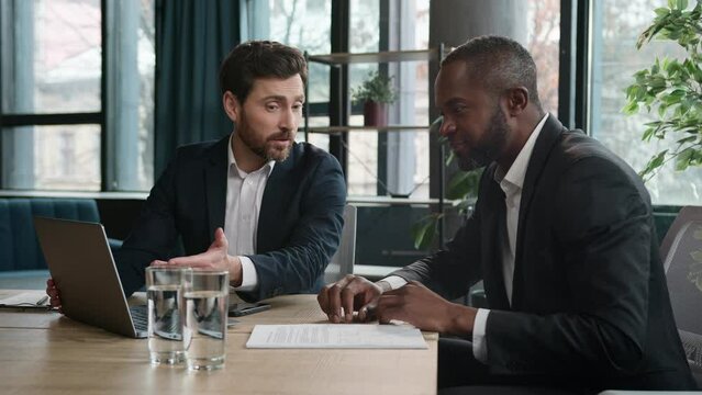 Diverse multiracial African American and Caucasian men colleagues coworkers businessmen financial agents discuss business project startup with laptop in office work with papers documents paperwork