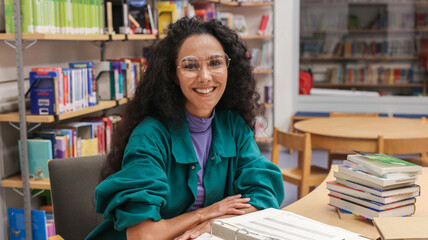 Portrait of a charming smiling Oriental girl with curly hair wearing glasses sitting at a school desk in a classroom or school library, backdrop of bookshelves smiling directly looking at the camera. - obrazy, fototapety, plakaty