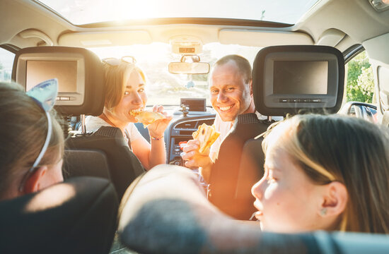 Young couple with daughters eating just cooked Italian pizza sitting in modern car with transparent roof. Happy family moments, childhood, fast food eating or auto journey lunch break concept image.