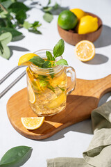 Lemonade drink or cold tea with fresh lemon, mint and ice in a jar. Refreshing citrus mojito cocktail on a wooden board on a  light background with fresh fruits and shadows.